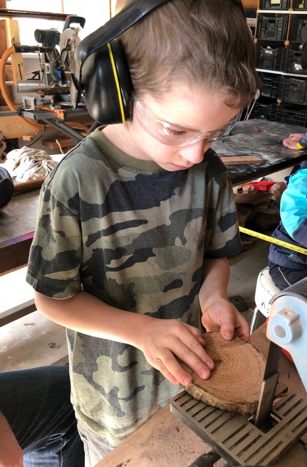 Boy wearing safety glasses and ear protection sanding a piece of wood on electric sander.